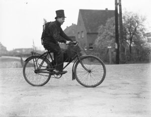 altes schwarzweißfoto von einem Schornsteinfeger auf Fahrrad, im Hintergrund ein älteres Haus