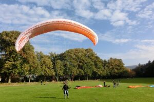 Gleitschirmlandung auf einer flachen grünen Wiese vor hohen Bäumen, bewölkter blauer Himmel