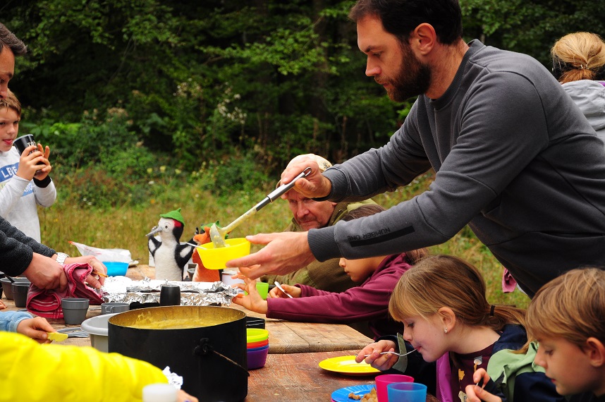 Outdoor-Kochen 2025_6 Kinder und Erwachsene beim gemeinsamen Essen im Wald auf einer Lichtung. Ein Mann schöpft gerade Kartoffelsuppe aus einem großen Topf