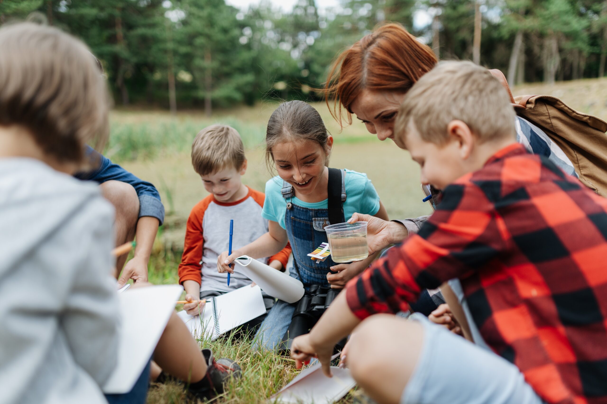 knieende Kinder und eine Frau entdecken etwas auf einer trockenen Wiese und notieren ihre Beobachtung