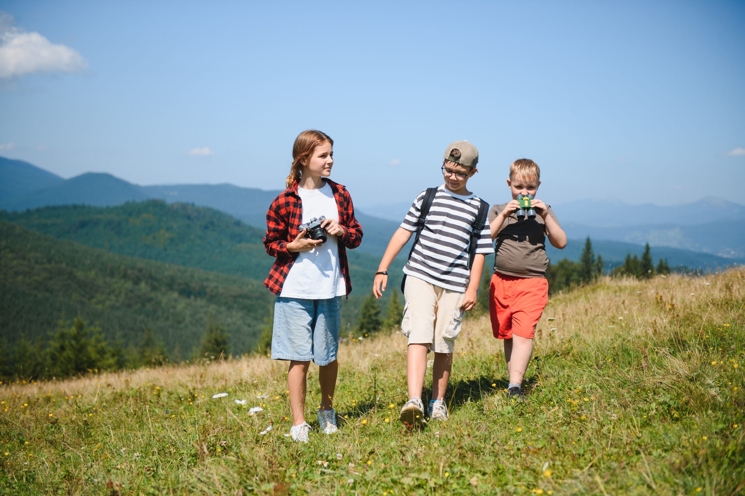 drei Kinder mit Fernglas und Fotoapparat wandern über eine Bergwiese