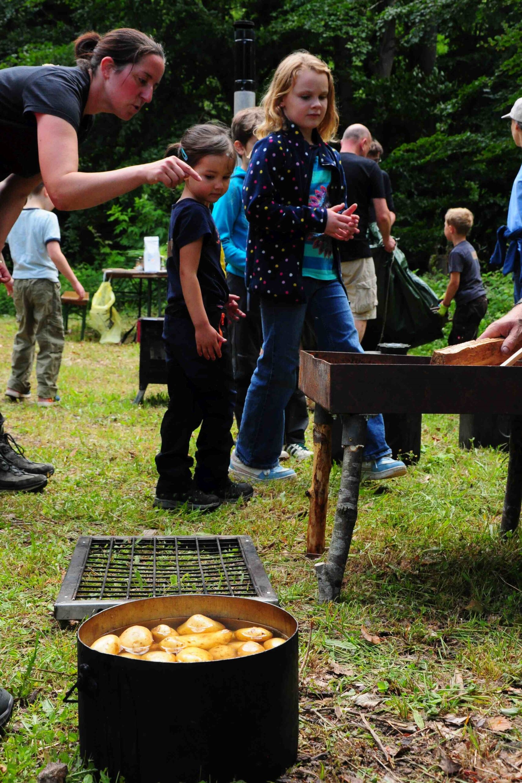 Kochstelle und ein Topf mit Kartoffeln in der Natur, Frau und Kinder beobachten sie