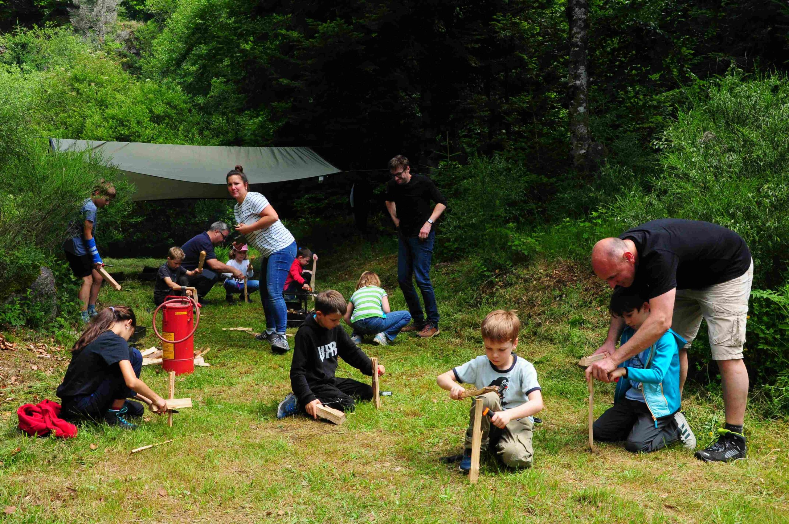 Kinder und Erwachsene auf einer Waldlichtung knieen oder stehen und spalten kleine Holzscheite