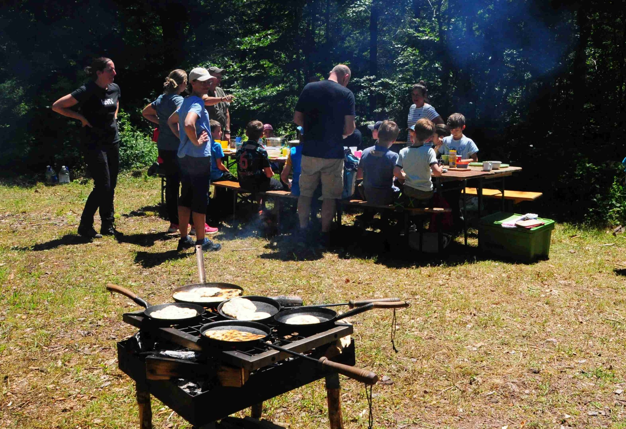 Kinder und Erwachsene auf einer Waldlichtung sitzen oder stehen um einen Tisch. Im Vordergrund braten Fladenbrote in Pfannen auf einer Kochstelle