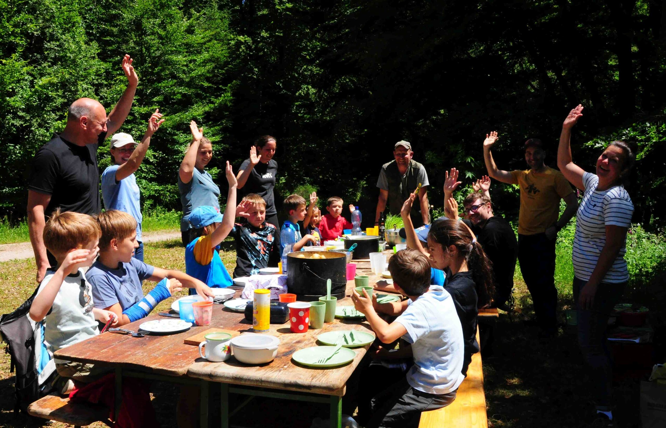 Kinder und Erwachsene auf einer Waldlichtung sitzen oder stehen um einen Tisch und geben ein freudiges Handzeichen