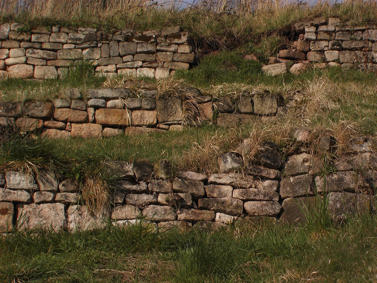 Trockenmauern aus unterschiedlich großen Steinen mit erkennbaren Spalten und Ritzen in drei Terrassen umgeben von Gräsern.