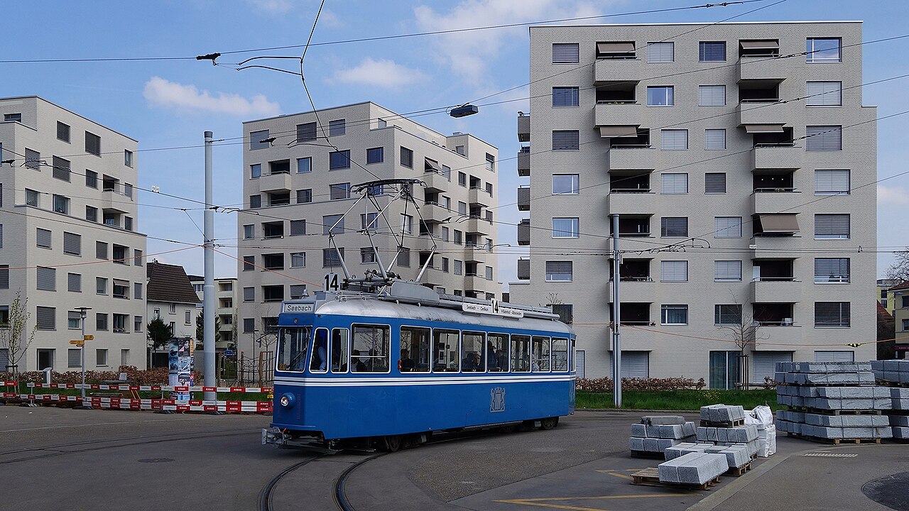 Eine blaue Straßenbahn vor drei großen Beton-Wohnhäusern, auf der rechten Seite verpacktes Baumaterial. Das Bild zeigt hauptsächlich versiegelte Fläche, die Hitze speichert.