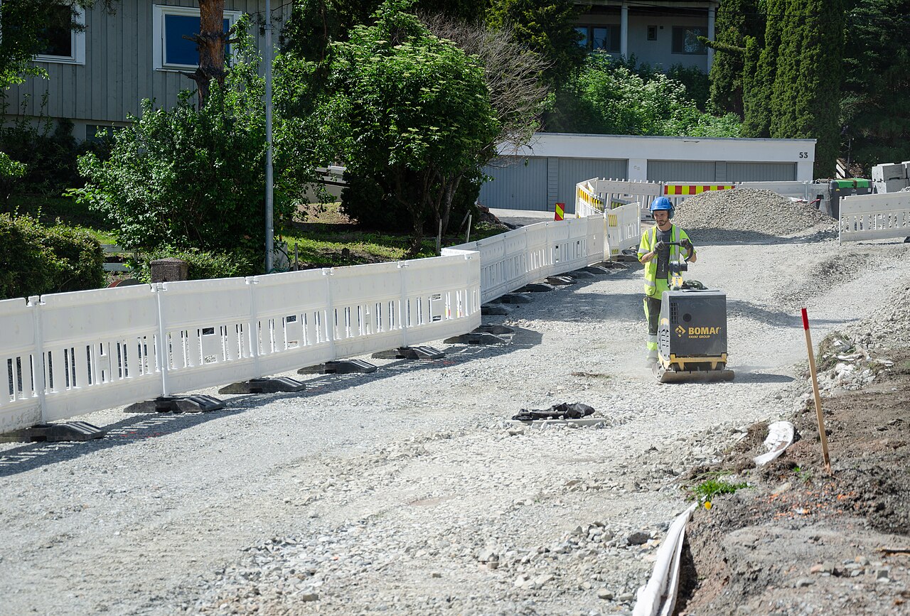 Foto einer Baustelle. Aktuell wird der Untergrund vorbereitet. Schotter und Kies sind bereits aufgebracht und werden von einem Arbeiter mit einem Rüttler verdichtet. Rüttler 