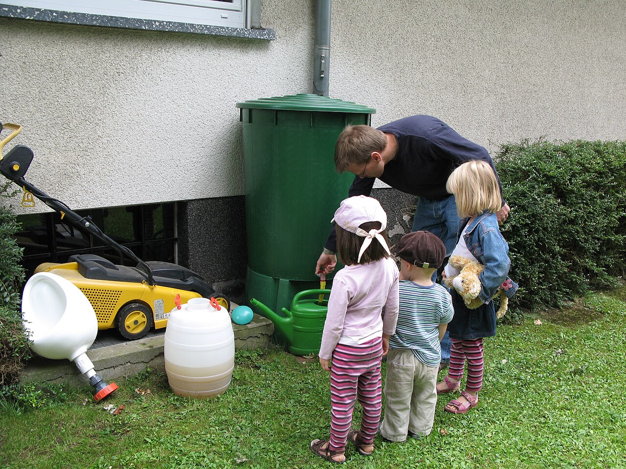Eine grüne Regentonne steht an einer Hauswand. Ein Mann füllt Wasser aus der Tonne in eine Gießkanne, drei Kinder schauen ihm dabei zu.