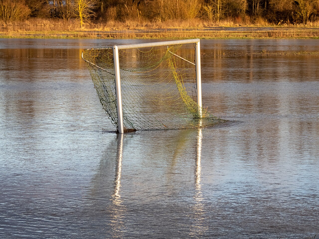 Ein Fußballtor steht auf einem mit Wasser überfluteten Platz. Im Hintergrund sieht man Bäume.