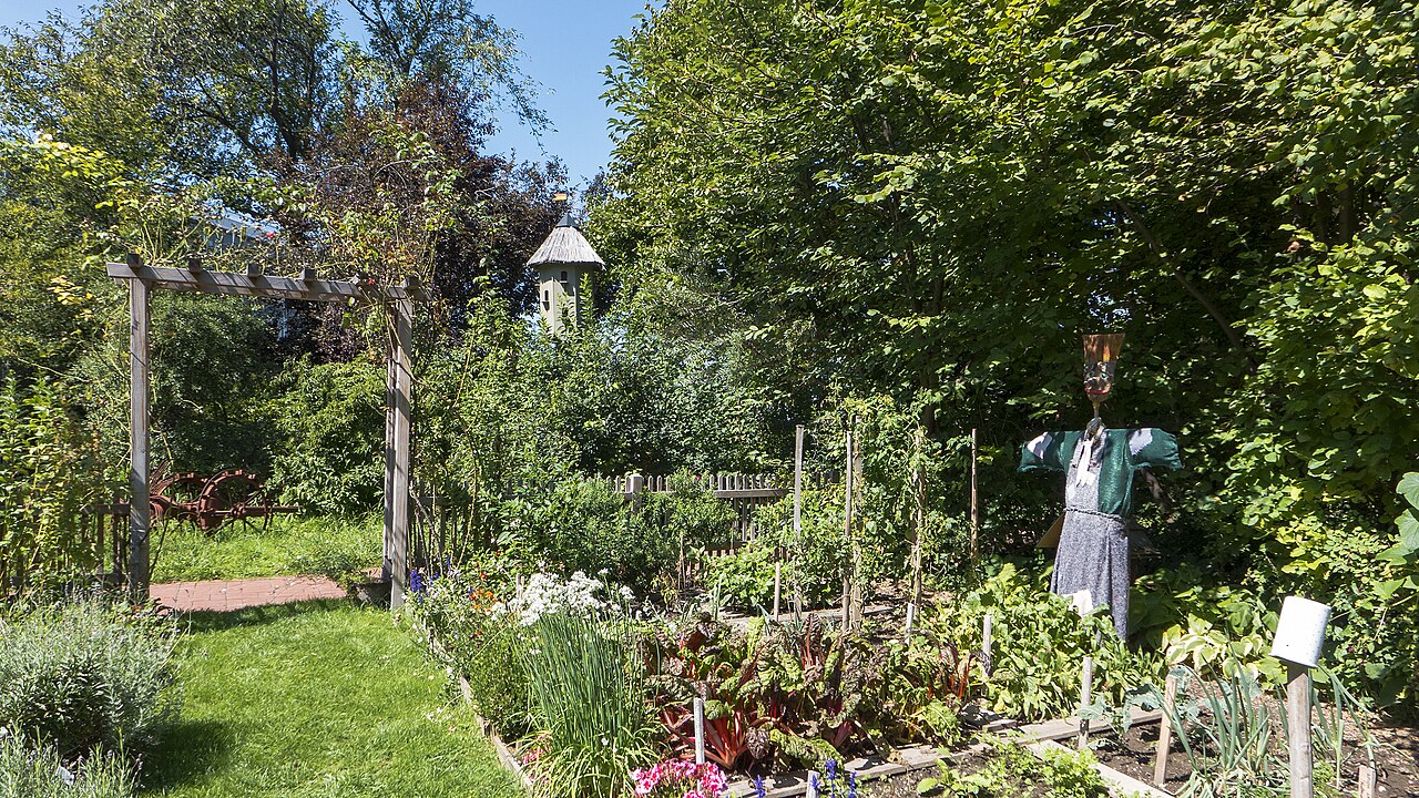 Ein sommerlicher Garten mit Gemüsebeet, in dem Tomaten, Mangold, Lauch und mehr wächst, außerdem Stauden, Sträucher und Bäume. Eine Vogelscheuche steht im Beet und ein kleines Holztor führt zu einem anderen Teil des Gartens.