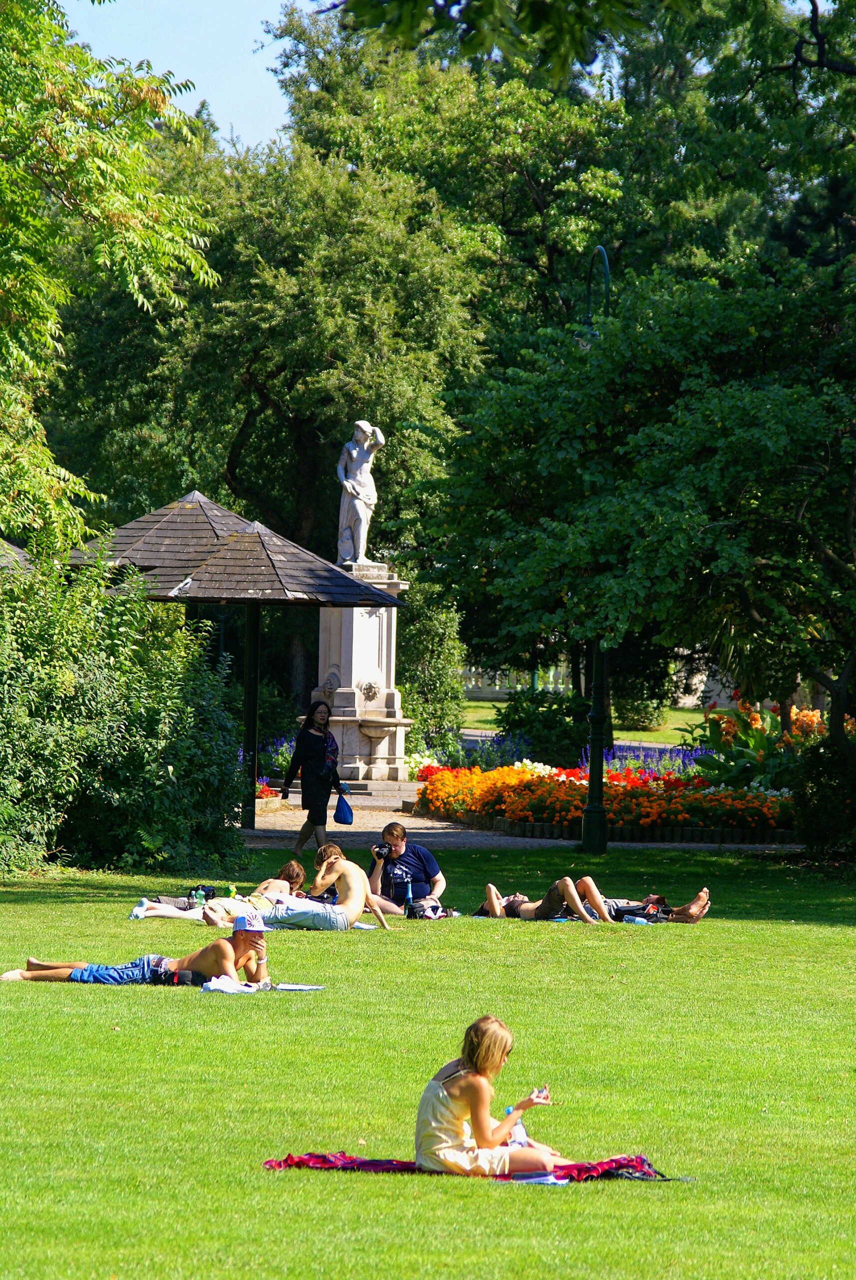 Wenn es warm wird, verbringen die Menschen gern Zeit im Grünen. Wie hier in einem Stadtpark. Menschen sitzen und liegen auf dem Rasen, im Hintergrund sind Bäume, eine Statue, Überdachungen und Blumenbeete.