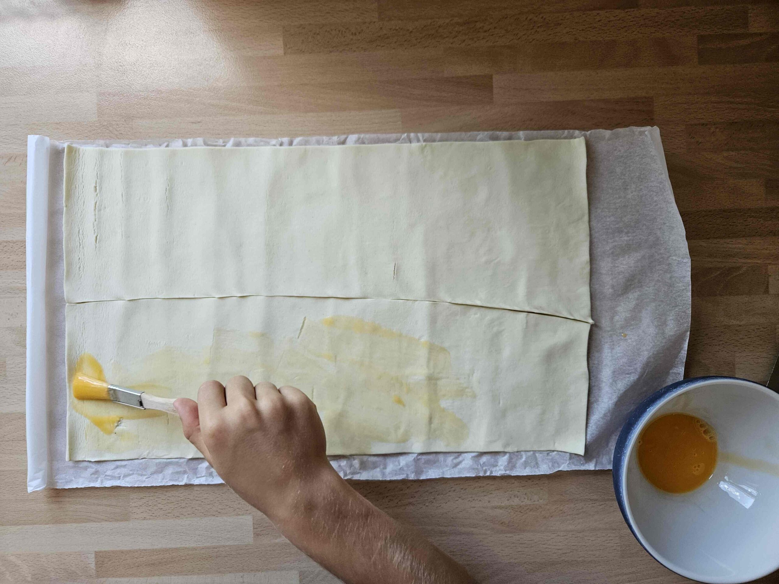 Ausgerollter Blätterteig, der mit Hilfe eines Pinsels Eigelb bestrichen wird.